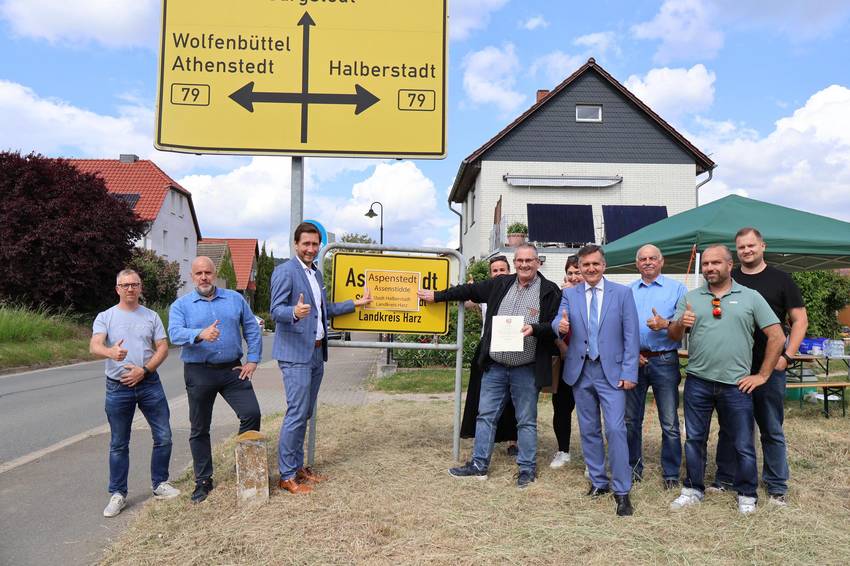 Mehrere Personen stehen vor dem Ortseingangsschild Aspenstedt. Halberstadts Oberbürgermeister Daniel Szarata hält zusammen mit Ortsbürgermeister Rüdiger Müller einen Ausdruck des neuen Ortschilds vor das aktuelle. Mehrere Personen stehen vor dem Ortseingangsschild Aspenstedt. Halberstadts Oberbürgermeister Daniel Szarata hält zusammen mit Ortsbürgermeister Rüdiger Müller einen Ausdruck des neuen Ortschilds vor das aktuelle.