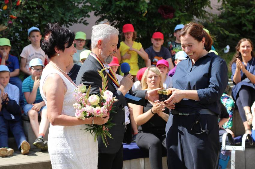 Dirk Clement übergibt seiner Nachfolgerin Veronika Berthold die Schulleiterin Stempel. Im Hintergrund sitzen Kinder mit Schirmmützen.