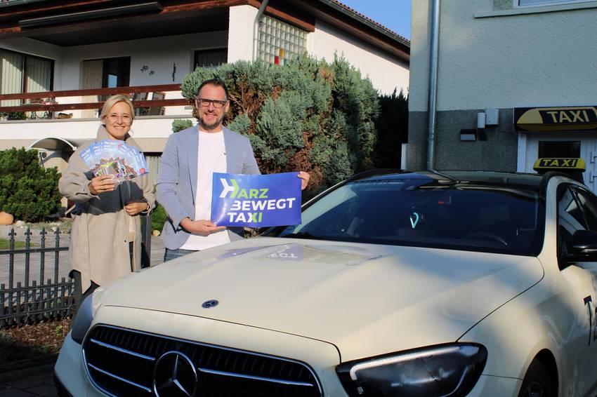 Zwei Personen stehen neben einem Taxi. Der Mann, der direkt neben dem Taxi steht, hält ein harzbewegt Schild in der Hand, die Frau daneben ein paar Flyer.