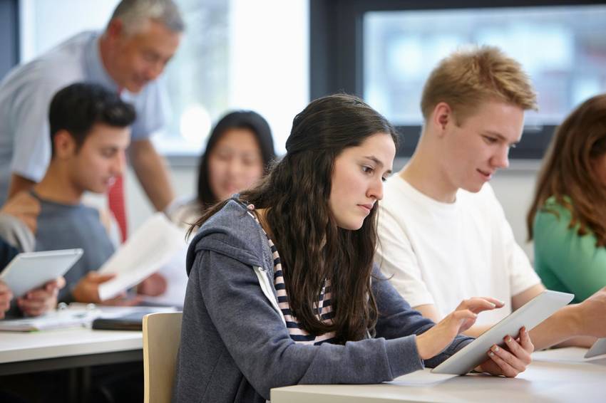 Unterrichtsituation. Im Vordergrund ist eine Schülerin am Tablet, daneben sitzt ein Schüler, im Hintergrund steht ein Lehrer, der etwas erklärt.