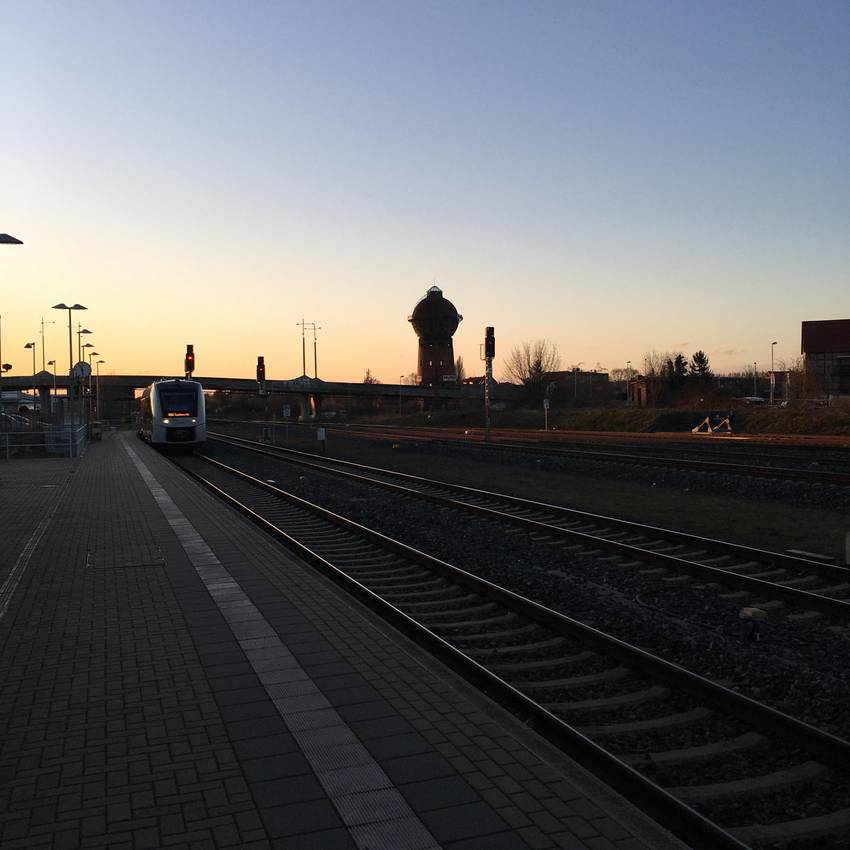 Der Halberstädter Bahnhof zum Sonnenuntergang. Ein Zug fährt ein, im Vordergrund sind die Gleise zu sehen, im Hintergrund der Wasserturm.