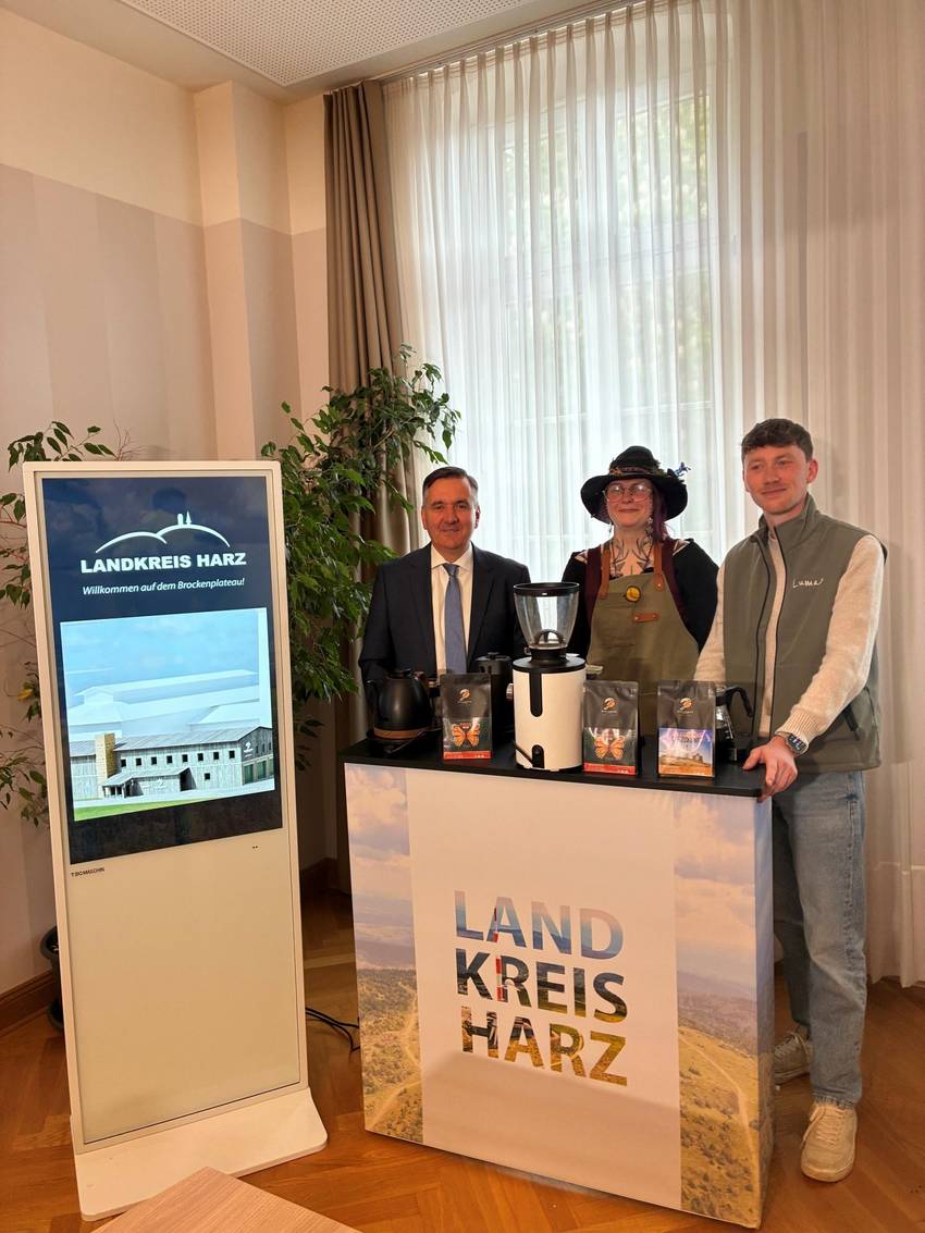 Gruppenfoto: v.l.n.r. Landrat Thomas Balcerowski, Lisa, die als Hexe verkleidet ist und Geschäftsführer Luran Engelhardt . Die drei Personen stehen hinter einem Landkreis Harz Tresen. Links neben dem Landrat steht eine Medienwand, die eine Animation der Kaffeerösterei auf dem Brocken zeigt. Gruppenfoto: v.l.n.r. Landrat Thomas Balcerowski, Lisa, die als Hexe verkleidet ist und Geschäftsführer Luran Engelhardt . Die drei Personen stehen hinter einem Landkreis Harz Tresen. Links neben dem Landrat steht eine Medienwand, die eine Animation der Kaffeerösterei auf dem Brocken zeigt.