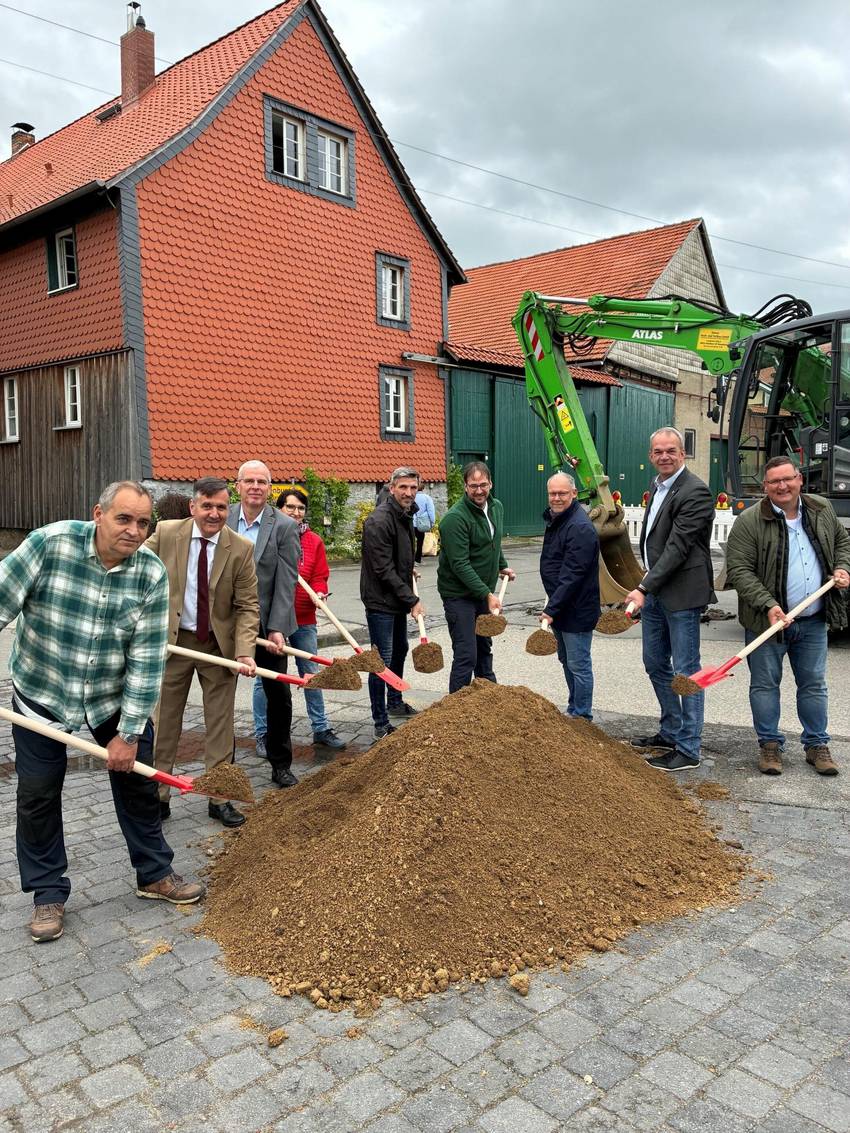 Gruppenfoto mit mehreren männlichen Perosnen, die einen Spaten in der Hand hlaten und hinter einem Sandberg stehen. Im Hintergrund sind Häuser und ein grüner Bagger.