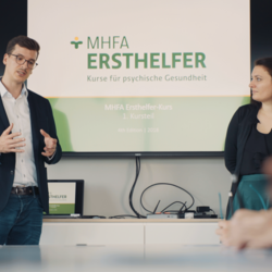 Zwei Menschen stehen vor einer Tafel mit dem Logo der Firma und schulen andere Menschen.