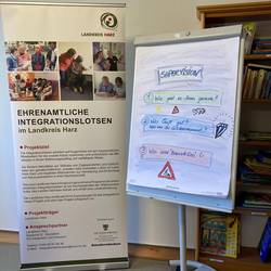 Ein Rollup der ehrenamtlichen Integrationslotsen. Daneben steht ein Flipchart mit der Aufschrift Supervision, darunter 1. Wie geht es Ihnen heute? 2. Was läuft gut? Was ist Ihr Glitzermoment? 3. Was sind "Baustellen"?