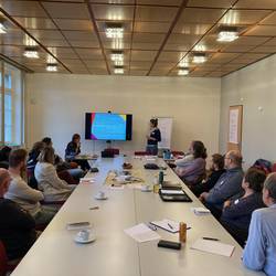 12 Personen sitzen an einem großen Tisch und schauen zu einer Präsentation, die von einer Dame gehalten wird.