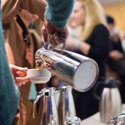 Café Miteinander Ein Tisch mit mehreren silbernen Kannen, vermutlich Kaffee. Eine Person, halb von hinten, die sich aus einer Kanne ein Getränk in die Tasse schüttet.
