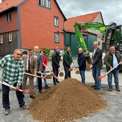 Gruppenfoto mit mehreren männlichen Perosnen, die einen Spaten in der Hand hlaten und hinter einem Sandberg stehen. Im Hintergrund sind Häuser und ein grüner Bagger. Gruppenfoto mit mehreren männlichen Perosnen, die einen Spaten in der Hand hlaten und hinter einem Sandberg stehen. Im Hintergrund sind Häuser und ein grüner Bagger.
