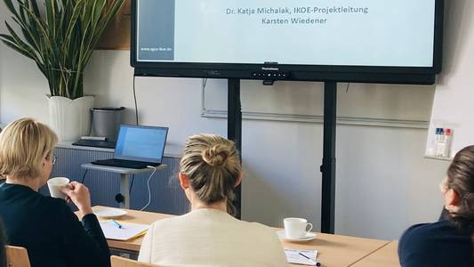 Mehrere Personen sitzen an einem Tisch und schauen auf einen großen Bildschirm, der eine Präsentation zeigt.