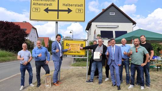 Mehrere Personen stehen vor dem Ortseingangsschild Aspenstedt. Halberstadts Oberbürgermeister Daniel Szarata hält zusammen mit Ortsbürgermeister Rüdiger Müller einen Ausdruck des neuen Ortschilds vor das aktuelle.