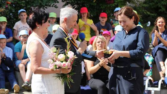 Dirk Clement übergibt seiner Nachfolgerin Veronika Berthold die Schulleiterin Stempel. Dirk Clement übergibt seiner Nachfolgerin Veronika Berthold die Schulleiterin Stempel. Im Hintergrund sitzen Kinder mit Schirmmützen.