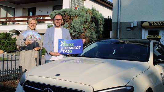 harzbewegt taxi Zwei Personen stehen neben einem Taxi. Der Mann, der direkt neben dem Taxi steht, hält ein harzbewegt Schild in der Hand, die Frau daneben ein paar Flyer.