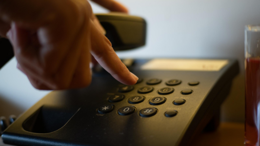 Eine Hand die auf einem Telefon eine Nummer tippt und den Hörer in der anderen Hand hat.