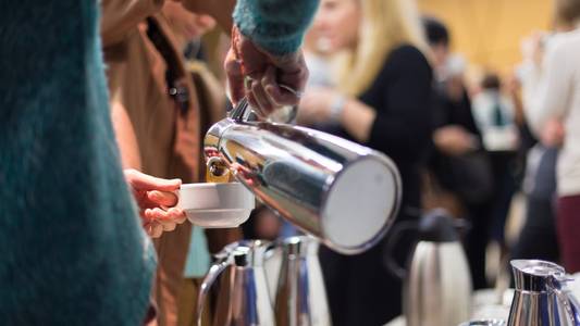 Ein Tisch mit mehreren silbernen Kannen, vermutlich Kaffee. Eine Person, halb von hinten, die sich aus einer Kanne ein Getränk in die Tasse schüttet.