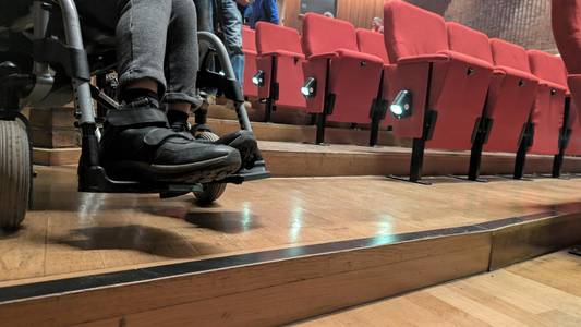 Eine Person im Rollstuhl steht im Zuschauerraum eines Theaters neben den Sitzreihen. Man sieht eine Stufe davor.
