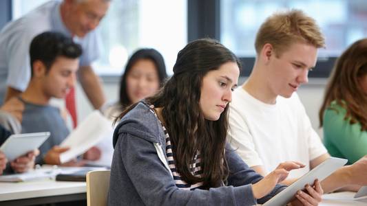 Unterrichtsituation. Im Vordergrund ist eine Schülerin am Tablet, daneben sitzt ein Schüler, im Hintergrund steht ein Lehrer, der etwas erklärt.