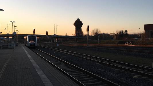 Der Halberstädter Bahnhof zum Sonnenuntergang. Ein Zug fährt ein, im Vordergrund sind die Gleise zu sehen, im Hintergrund der Wasserturm.