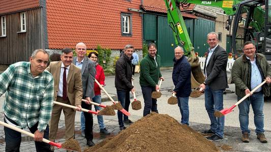 Gruppenfoto mit mehreren männlichen Personen, die einen Spaten in der Hand halten und hinter einem Sandberg stehen. Im Hintergrund ein Haus und ein grüner Bagger.
