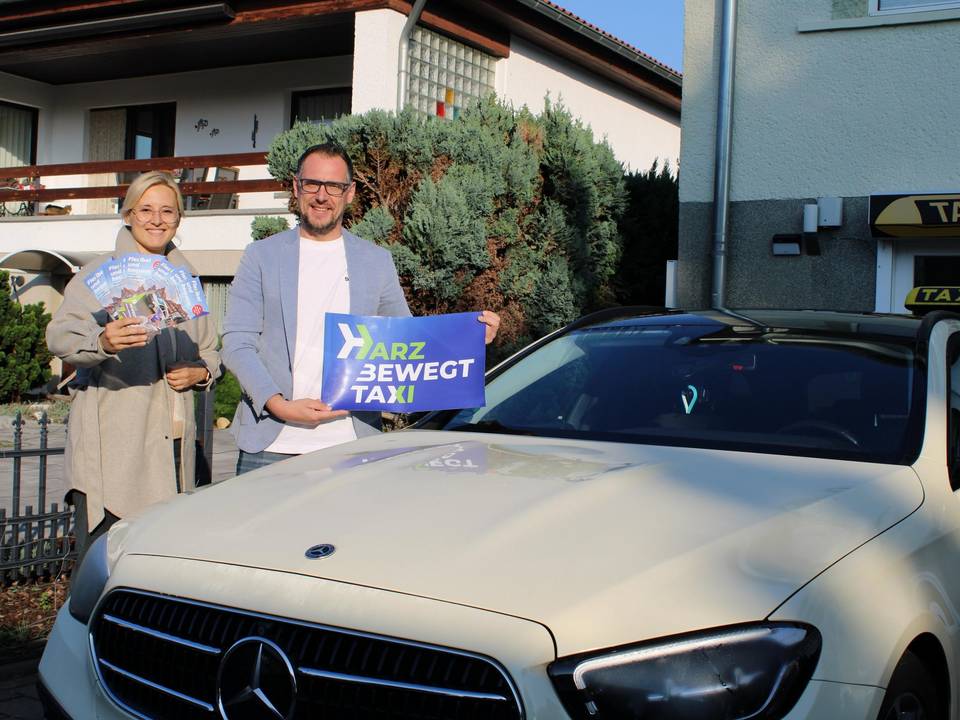 harzbewegt taxi Zwei Personen stehen neben einem Taxi. Der Mann, der direkt neben dem Taxi steht, hält ein harzbewegt Schild in der Hand, die Frau daneben ein paar Flyer.