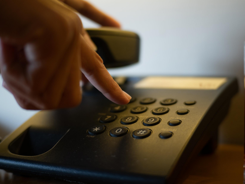 Eine Hand die auf einem Telefon eine Nummer tippt und den Hörer in der anderen Hand hat.