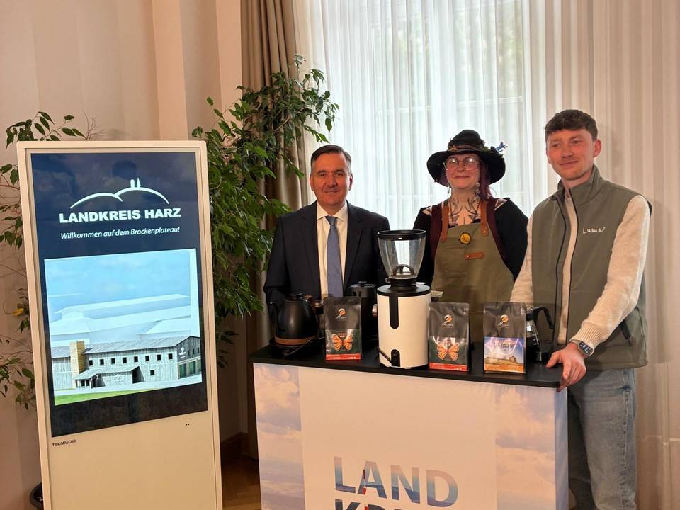 Gruppenfoto: v.l.n.r. Landrat Thomas Balcerowski, Lisa, die als Hexe verkleidet ist und Geschäftsführer Luran Engelhardt . Die drei Personen stehen hinter einem Landkreis Harz Tresen. Links neben dem Landrat steht eine Medienwand, die eine Animation der Kaffeerösterei auf dem Brocken zeigt. Gruppenfoto: v.l.n.r. Landrat Thomas Balcerowski, Lisa, die als Hexe verkleidet ist und Geschäftsführer Luran Engelhardt . Die drei Personen stehen hinter einem Landkreis Harz Tresen. Links neben dem Landrat steht eine Medienwand, die eine Animation der Kaffeerösterei auf dem Brocken zeigt.
