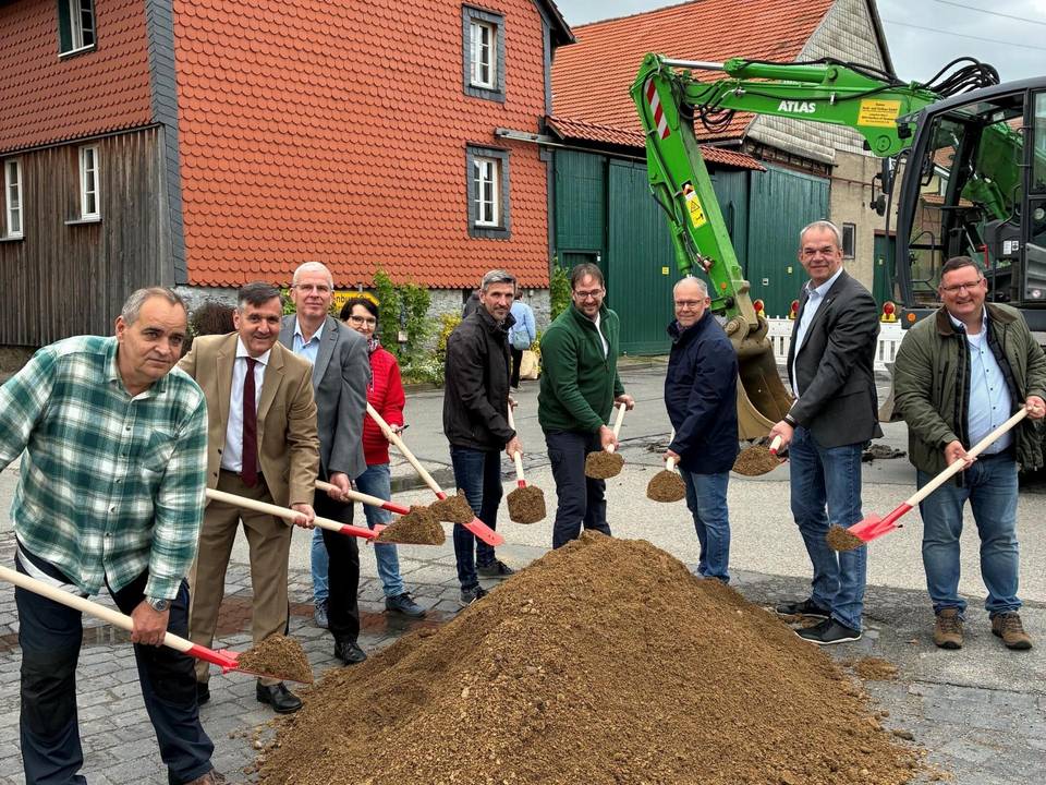 Gruppenfoto mit mehreren männlichen Perosnen, die einen Spaten in der Hand hlaten und hinter einem Sandberg stehen. Im Hintergrund sind Häuser und ein grüner Bagger.